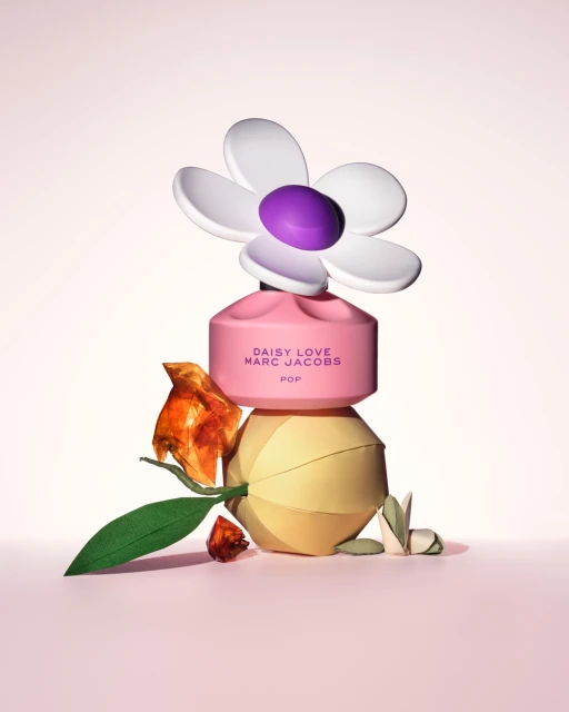 Pink Marc Jacobs Daisy Love POP perfume bottle with white daisy cap, paper flower, and leaves on a light pink background. 