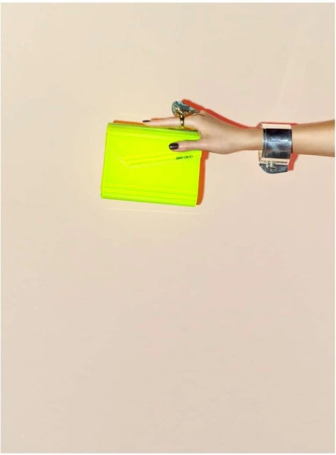 Hand holding a bright yellow clutch against a neutral background, accessorized with a cuff bracelet and a large statement ring.