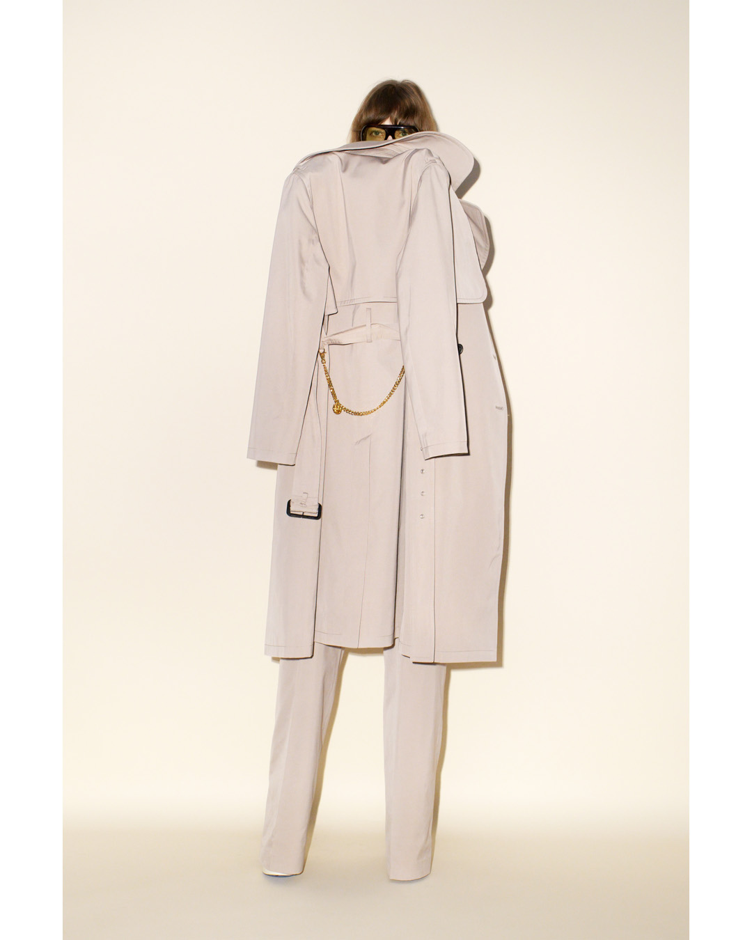 Back view of an adult female in a long beige trench coat with a gold chain, matching suit pants, and dark hair, indoors against an off-white backdrop.