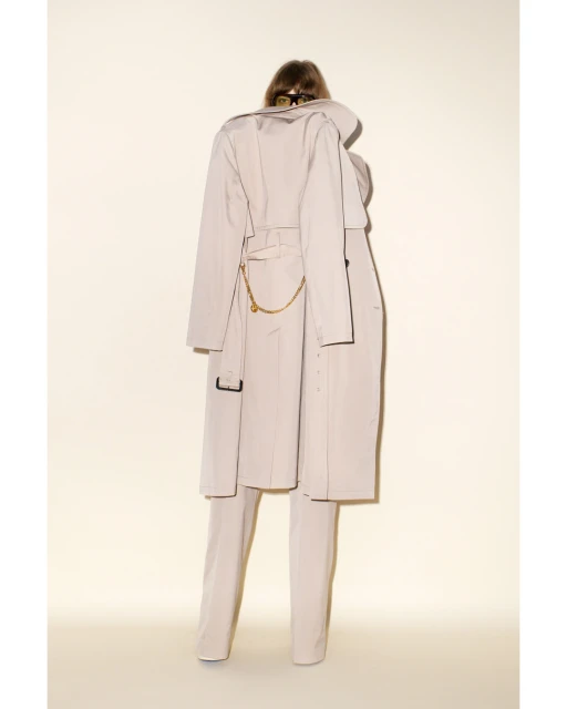 Back view of an adult female in a long beige trench coat with a gold chain, matching suit pants, and dark hair, indoors against an off-white backdrop.