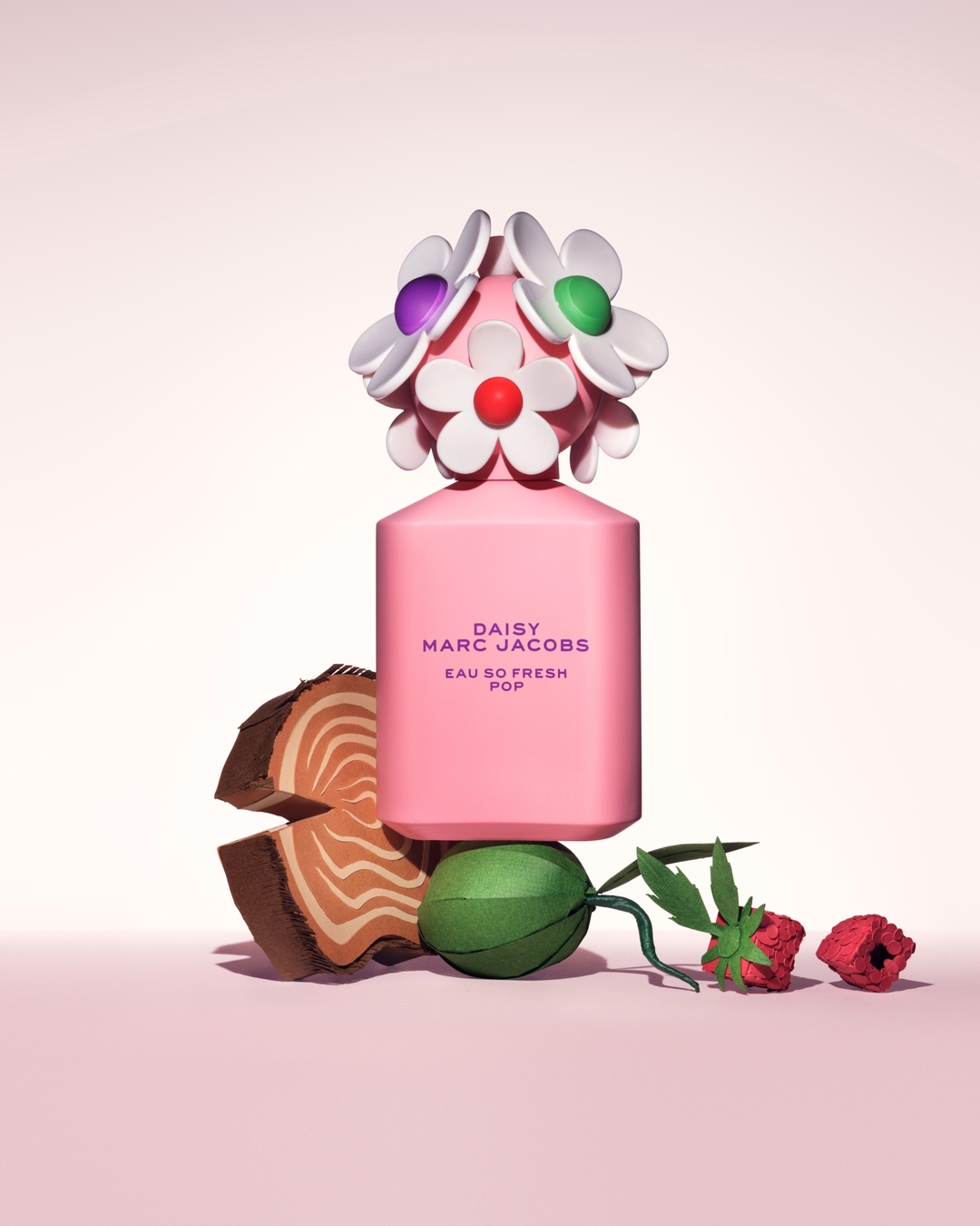 Pink Daisy Marc Jacobs perfume bottle with flower-shaped cap sits among cut wood, green paper, and red paper flowers on a pink backdrop.
