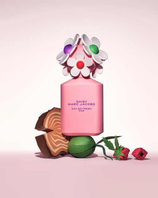 Pink Daisy Marc Jacobs perfume bottle with flower-shaped cap sits among cut wood, green paper, and red paper flowers on a pink backdrop.