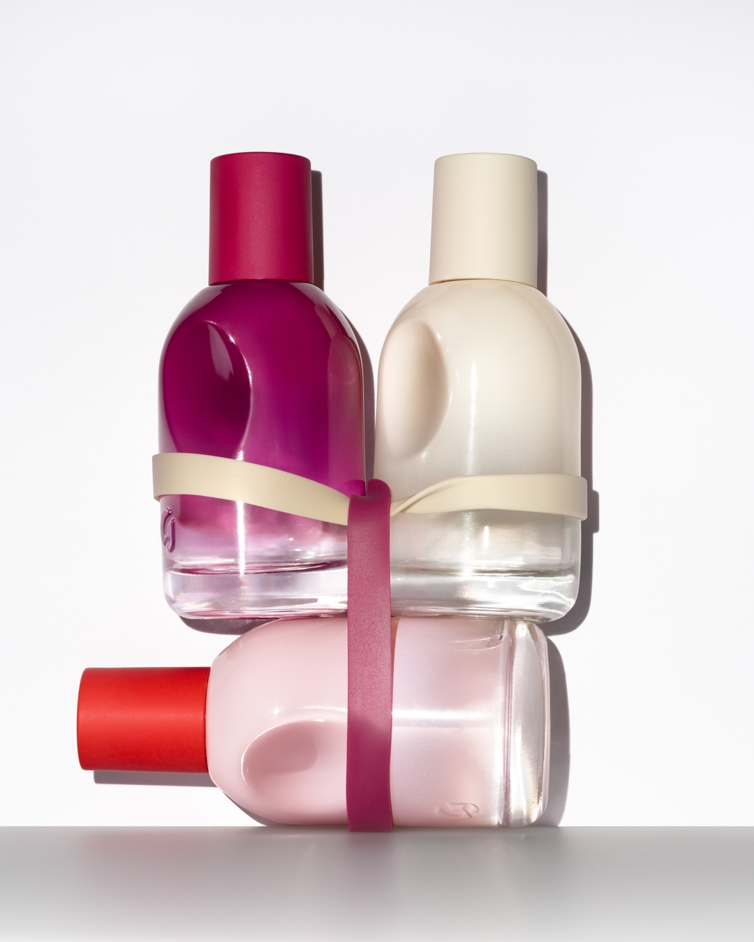 Three perfume bottles, varying in pink and red hues, are stacked and secured with light-colored bands against a bright white backdrop.