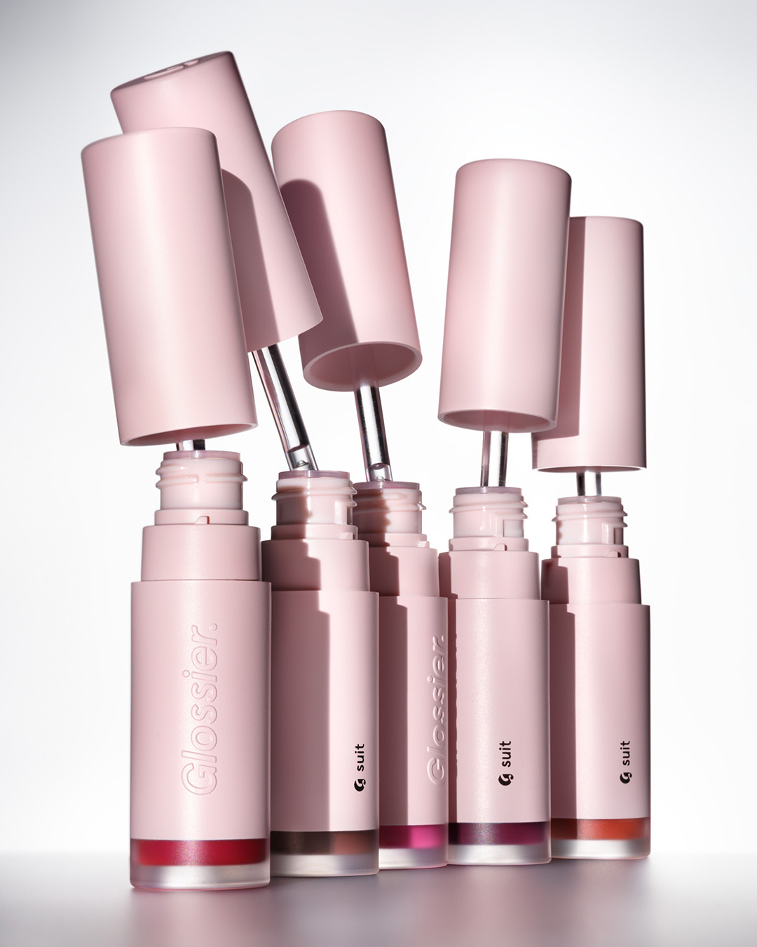 Several pink Glossier Gen G lipsticks stand upright against a white background, some with the caps removed showcasing the applicator wands.