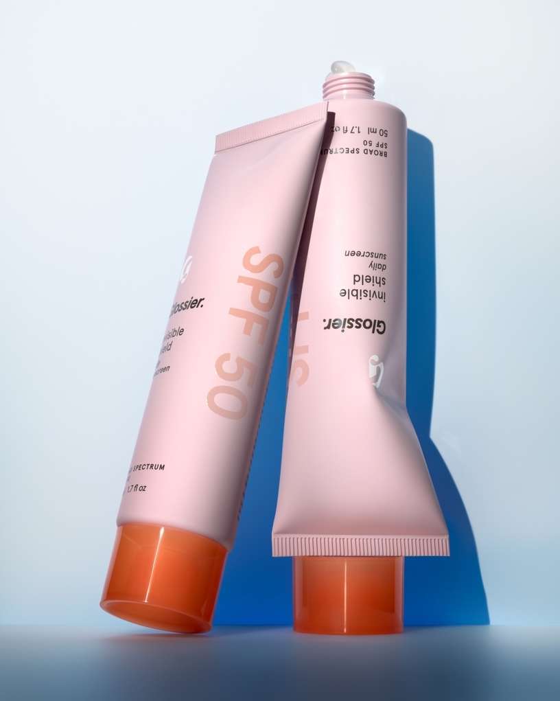Two pink tubes of Glossier Invisible Shield sunscreen with orange caps on a light blue background in a product shot.