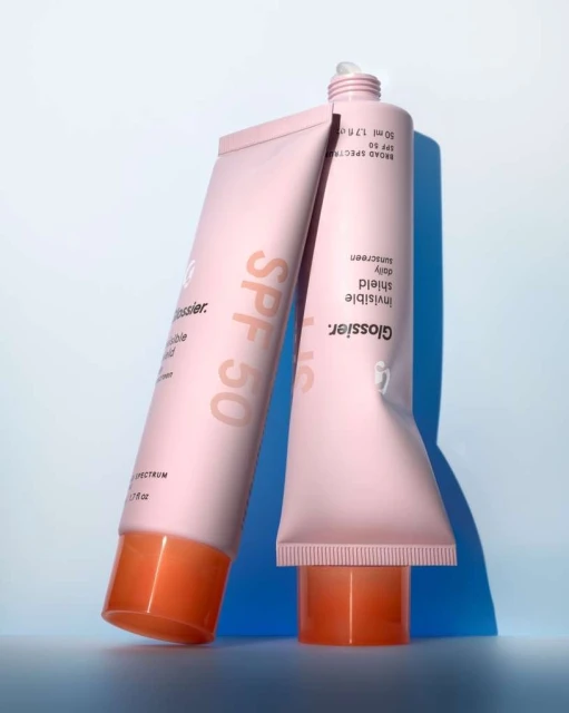 Two pink tubes of Glossier Invisible Shield sunscreen with orange caps on a light blue background in a product shot.