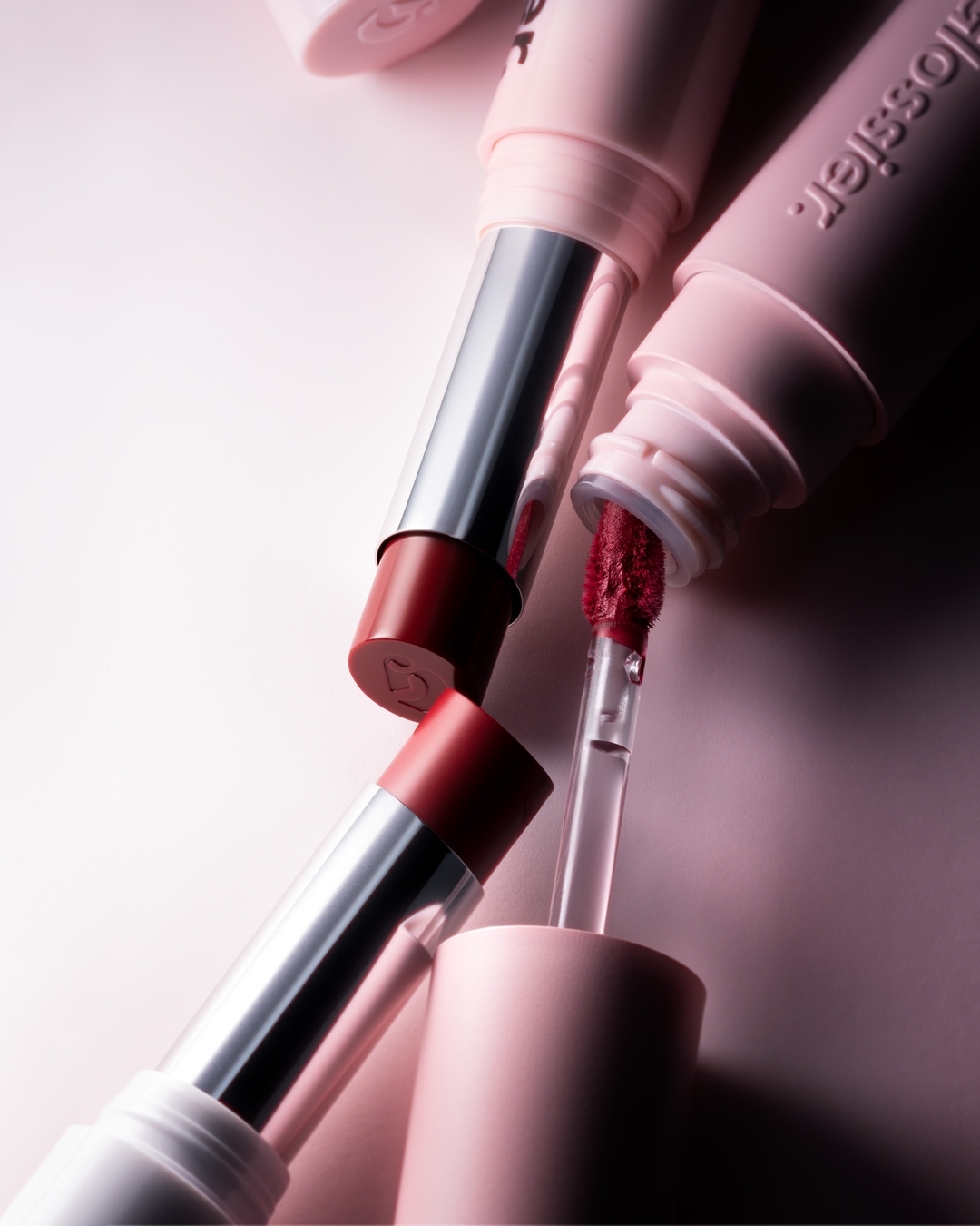 Close-up of Glossier lipsticks and lip gloss in a minimalist setting, showcasing the brand's makeup range with soft, diffused lighting.