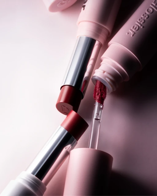 Close-up of Glossier lipsticks and lip gloss in a minimalist setting, showcasing the brand's makeup range with soft, diffused lighting.