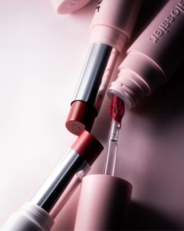 Close-up of Glossier lipsticks and lip gloss in a minimalist setting, showcasing the brand's makeup range with soft, diffused lighting.