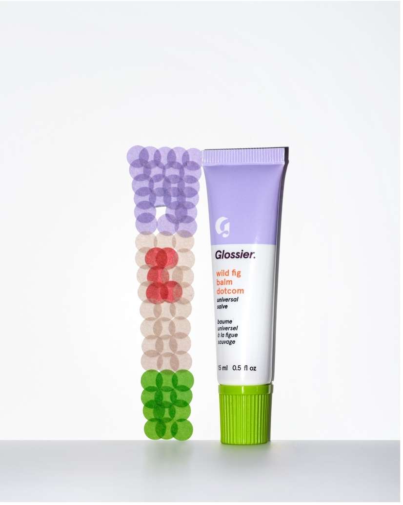 Cosmetic tube of Glossier balm in lilac and green packaging on white surface with blurred circles in purple, red and green.