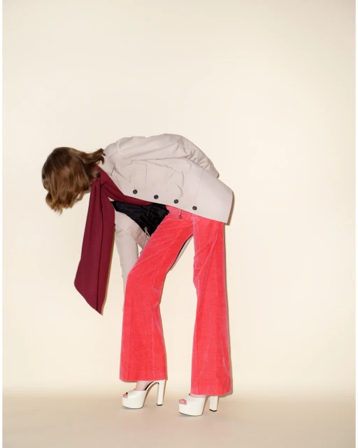 Fashionable woman in a beige coat with a red scarf and red flared pants bends over in a studio with a light beige background.