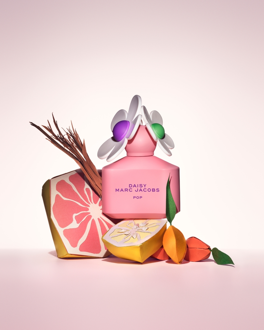 Marc Jacobs Daisy POP fragrance bottle stands out amidst artful paper citrus fruits, set against a soft pink backdrop.
