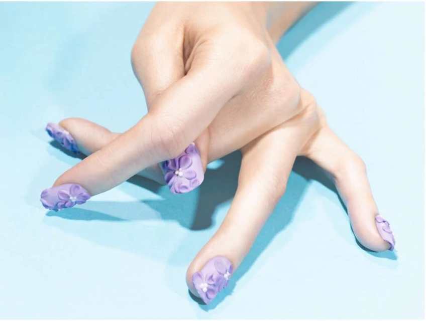 Close-up of a woman's hand with a lilac manicure adorned with floral nail art, set against a light blue background.