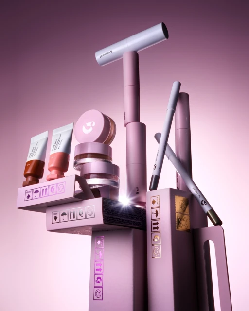 Cosmetic products are stacked on boxes, featuring tubes, jars, and pencils in pastel hues, against a soft pink gradient background.