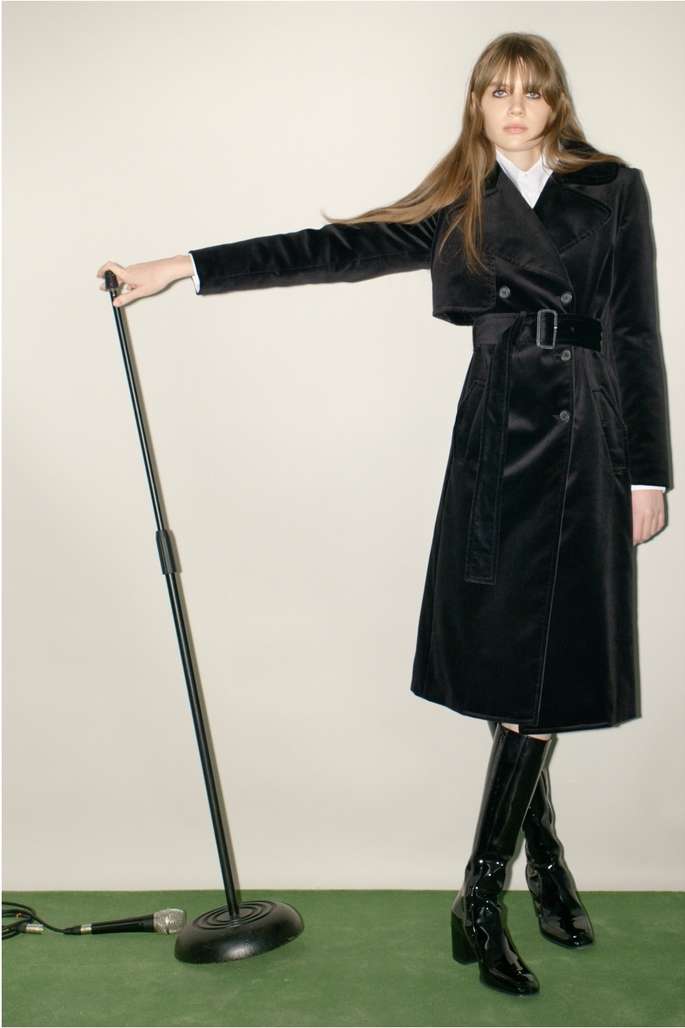 A woman in a black velvet trench coat poses in a studio with a microphone stand, showcasing a blend of fashion and retro stage elements.