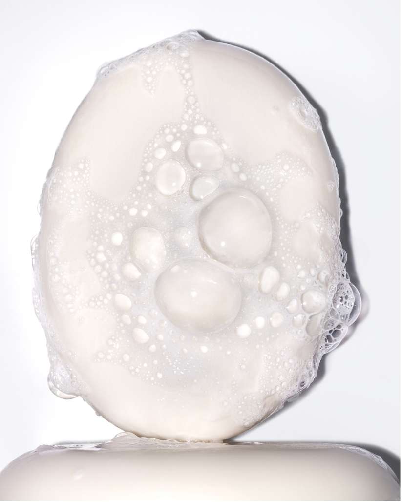 Close-up of an oval bar of soap with bubbles on top, sitting on another bar of soap, set against a clean, white background.
