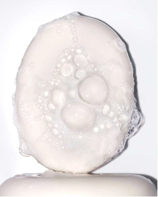 Close-up of an oval bar of soap with bubbles on top, sitting on another bar of soap, set against a clean, white background.