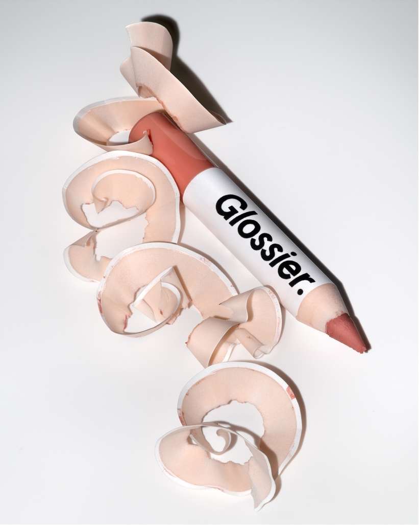 Close-up of a Glossier lip pencil with a peach-colored tip and white barrel, surrounded by peach-colored pencil shavings on white.