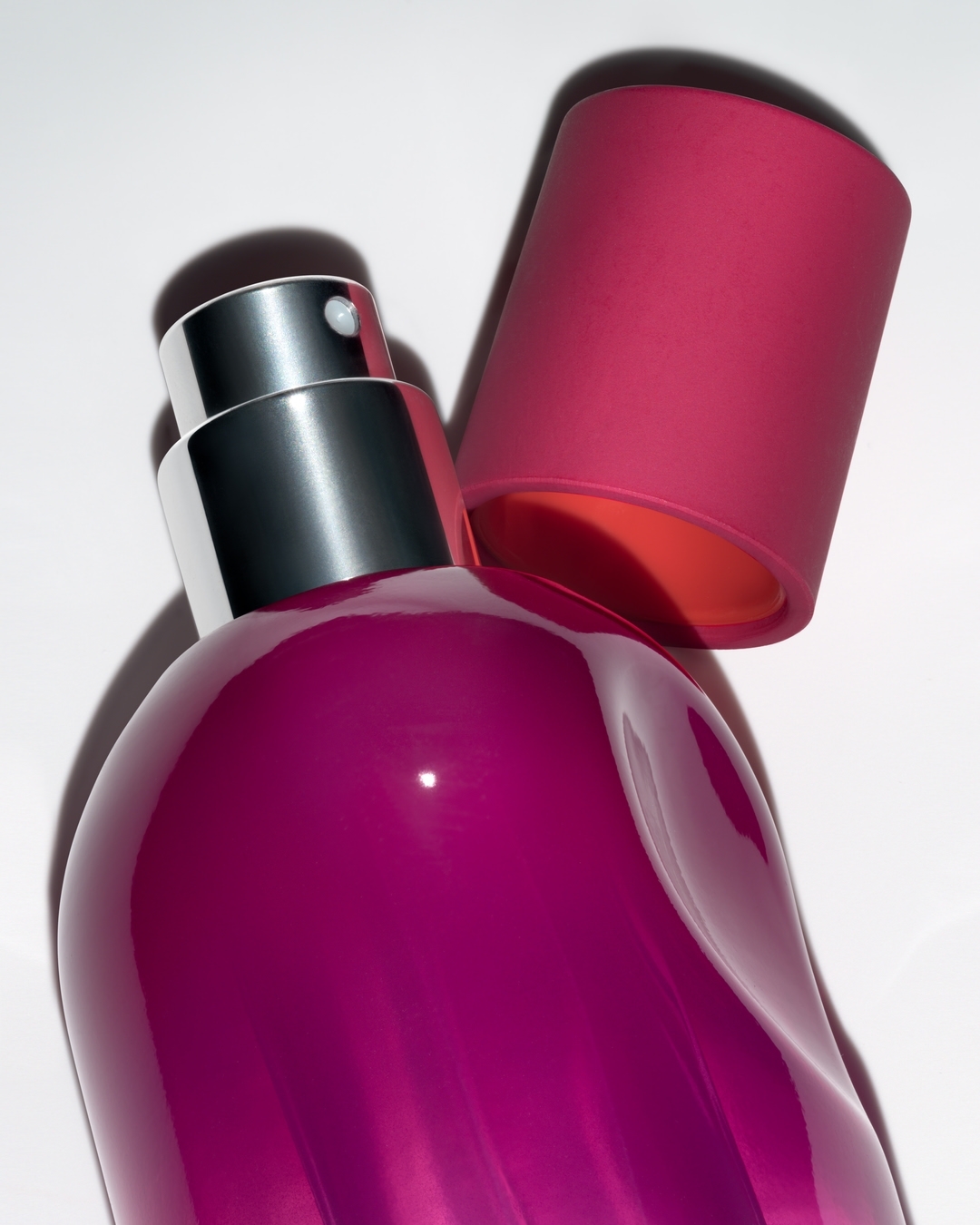 Close-up studio shot of a magenta bottle with a silver spray nozzle and a magenta lid on a white surface, casting a soft shadow.