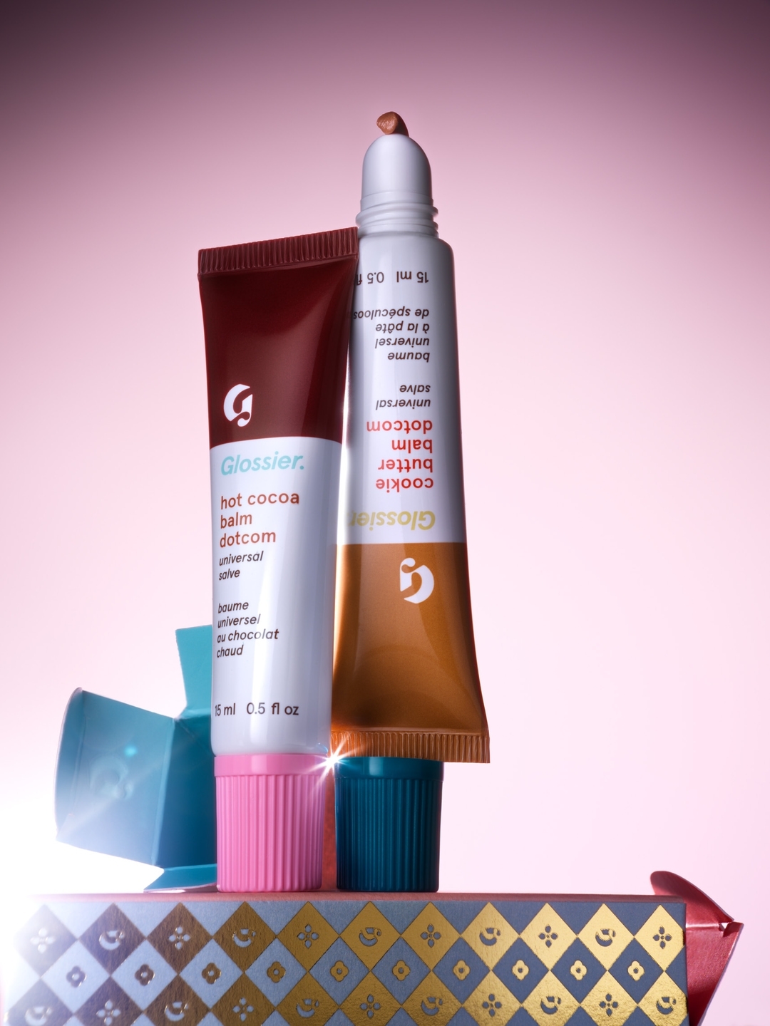 Two tubes of Glossier balm, "hot cocoa" and "cookie butter", stand on a patterned gift box against a pink gradient background.