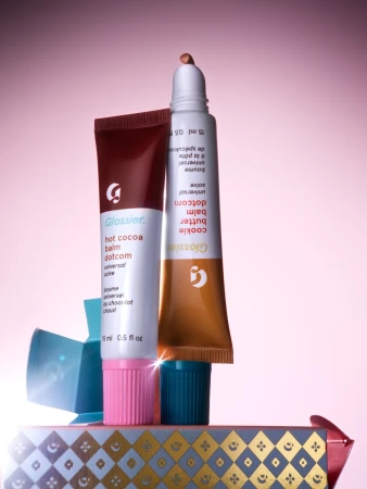 Two tubes of Glossier balm, "hot cocoa" and "cookie butter", stand on a patterned gift box against a pink gradient background.