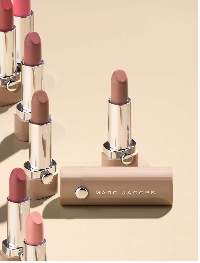 Several Marc Jacobs lipsticks in a row on a tan surface, some standing up, one laying down, showcasing colors and packaging.