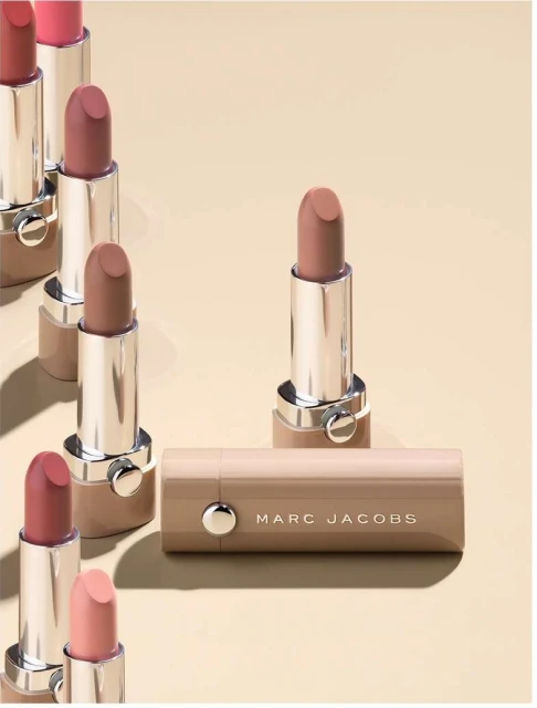 Several Marc Jacobs lipsticks in a row on a tan surface, some standing up, one laying down, showcasing colors and packaging.