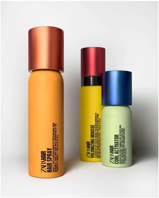 Three colorful ZARA hair product bottles in a studio setting: hair spray (orange), volumizing mousse (yellow), and curl activator (green).