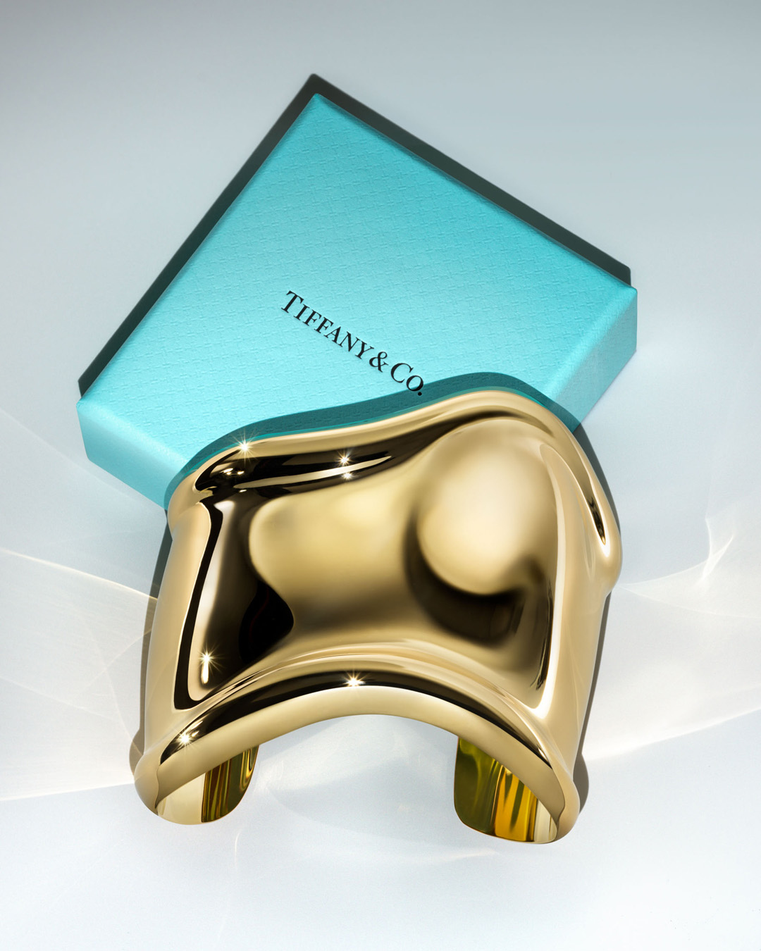 Gold metal cuff bracelet with a glossy finish, reflecting light, presented next to a light blue Tiffany & Co. branded box on white.