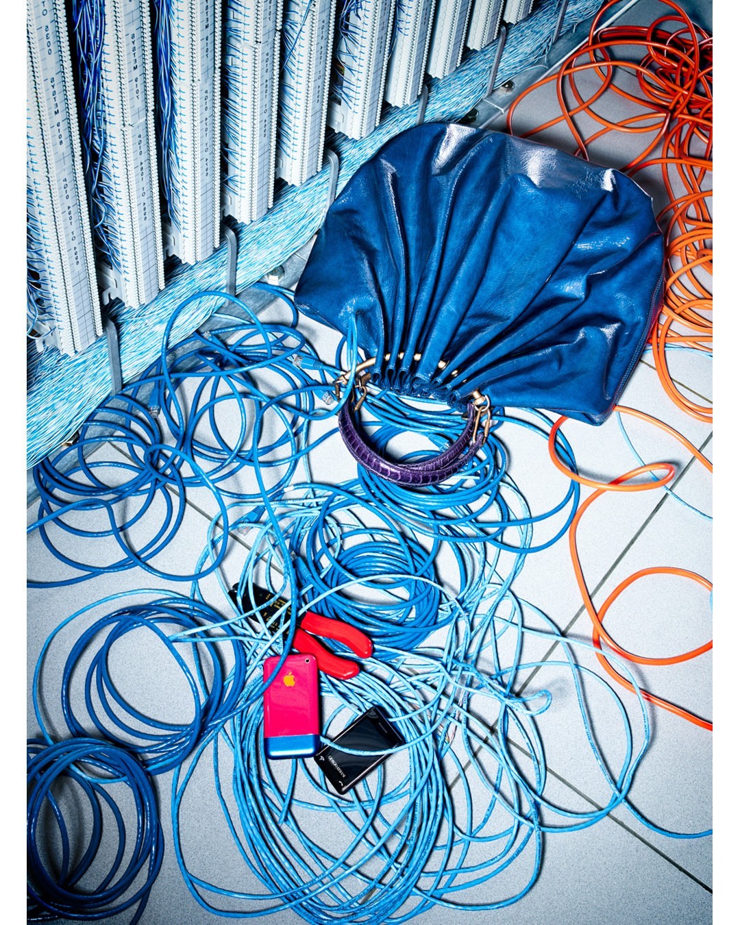 Fashion still life featuring a blue leather handbag with gold hardware, surrounded by a tangle of blue and orange computer cables.