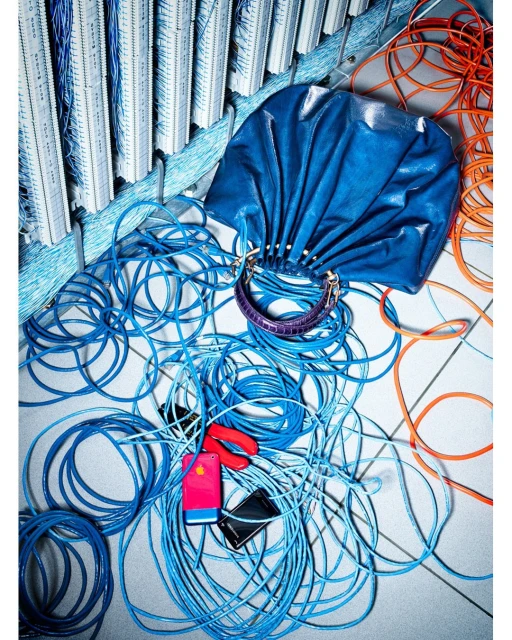 Fashion still life featuring a blue leather handbag with gold hardware, surrounded by a tangle of blue and orange computer cables.