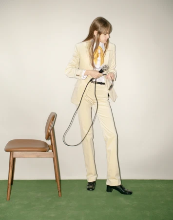 A woman in a cream corduroy suit holding a microphone stands next to a wooden chair on a green carpet.