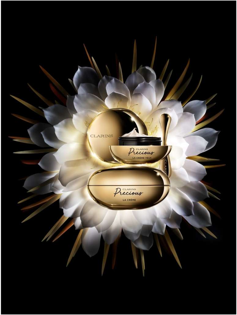 Three gold-toned Clarins Precious La Creme skincare containers arranged with a white, radial flower on a black background.