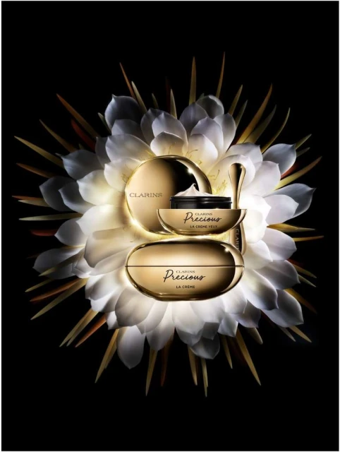 Three gold-toned Clarins Precious La Creme skincare containers arranged with a white, radial flower on a black background.
