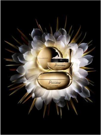 Three gold-toned Clarins Precious La Creme skincare containers arranged with a white, radial flower on a black background.