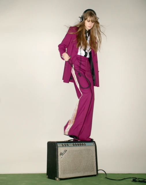 A young woman in a magenta suit and headphones stands on a vintage speaker against a neutral background, exuding a lively and stylish vibe.
