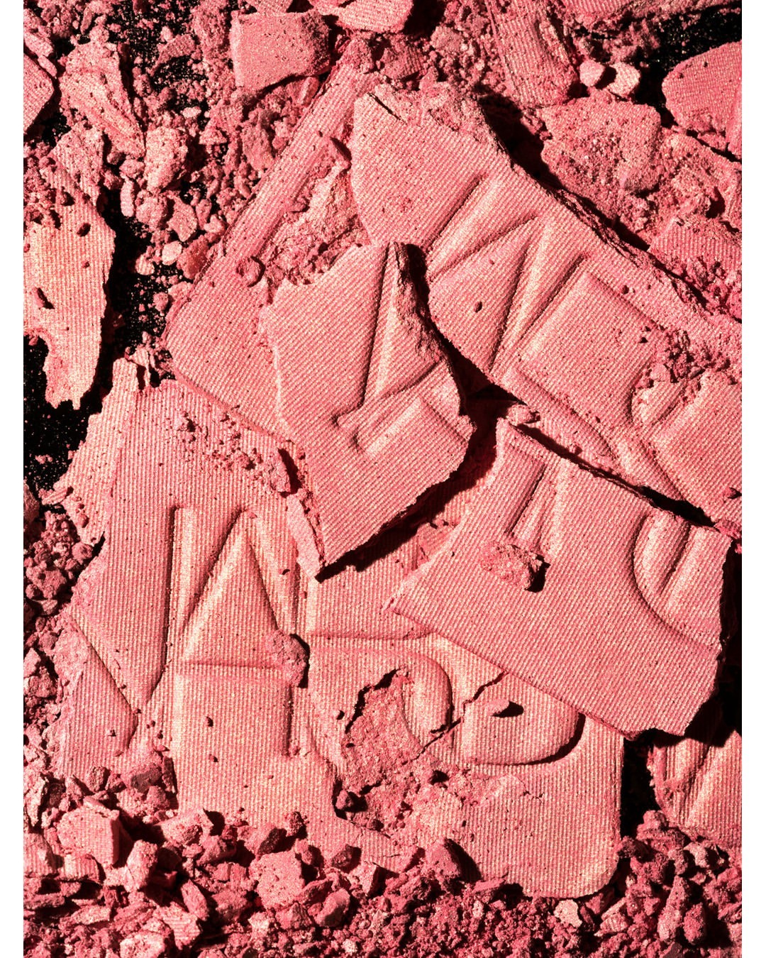 Close-up of broken rose-pink NARS blush powder showing crushed texture, product lettering, and coloration against a dark backdrop.