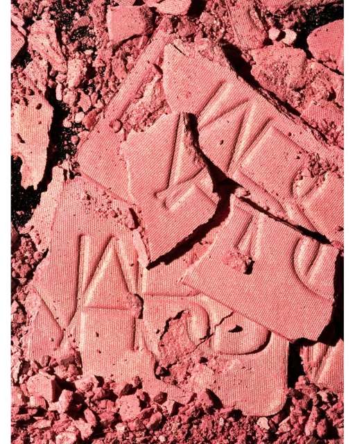 Close-up of broken rose-pink NARS blush powder showing crushed texture, product lettering, and coloration against a dark backdrop.