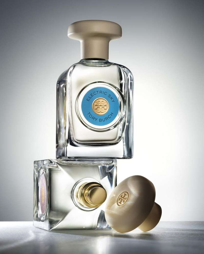 Two stacked Tory Burch Electric Sky perfume bottles with a cap lying beside them in a studio with white and grey gradient background.