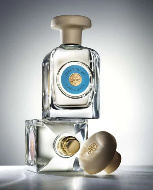Two stacked Tory Burch Electric Sky perfume bottles with a cap lying beside them in a studio with white and grey gradient background.