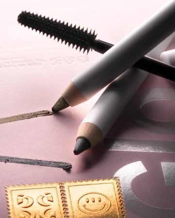 Close-up still life of eye makeup pencils and mascara wand on a pink surface, emphasizing beauty product textures and cosmetic application.