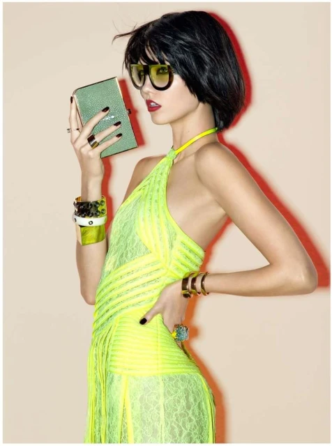A fashionable woman with short dark hair in a neon yellow dress holds a small, pale green box in a studio setting.