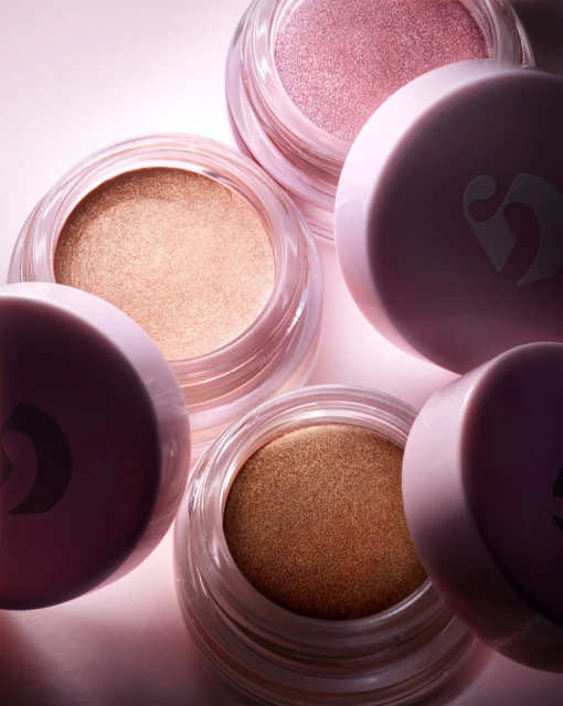 Close-up of Glossier eye shadow pots in pink and bronze tones with clear containers and pink lids on a pink background.