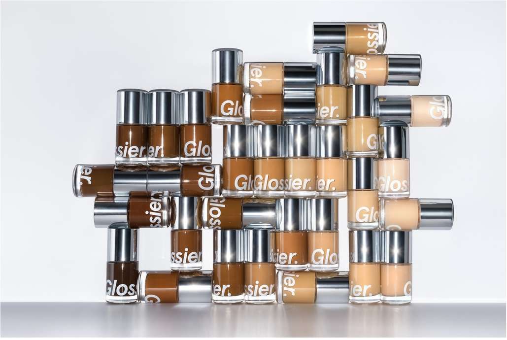 Multiple bottles of Glossier foundation in various shades stacked in front of a white backdrop in a studio-lit beauty product shot.