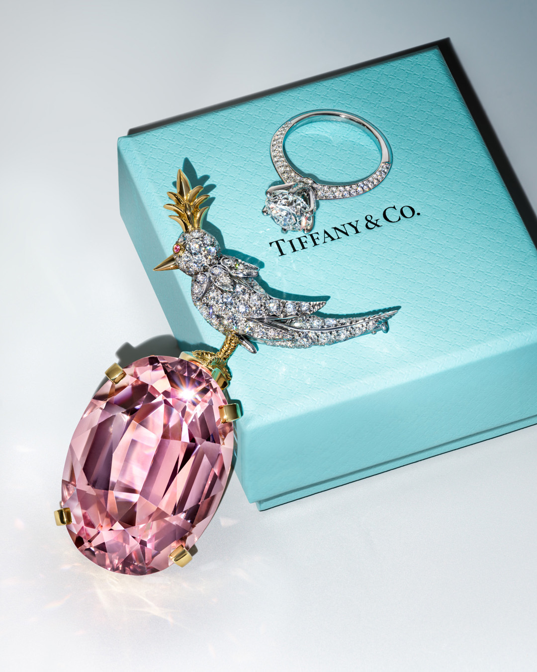 Close-up of a bird-shaped brooch with a large pink gemstone, a diamond ring, and a Tiffany & Co. jewelry box, luxurious and elegant.