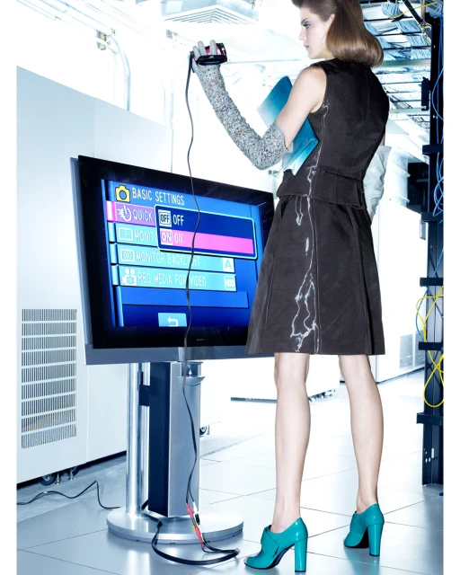 Woman in dress holds device connected to flat screen TV in a brightly lit room with white walls and networking cables.