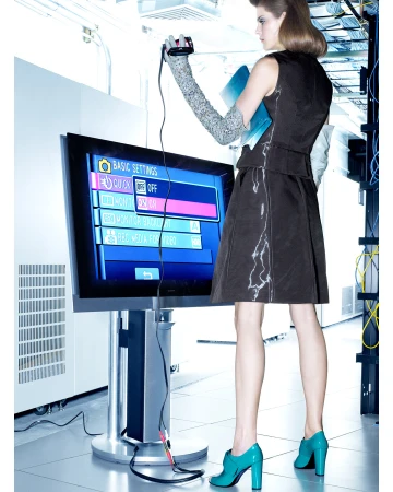 Woman in dress holds device connected to flat screen TV in a brightly lit room with white walls and networking cables.