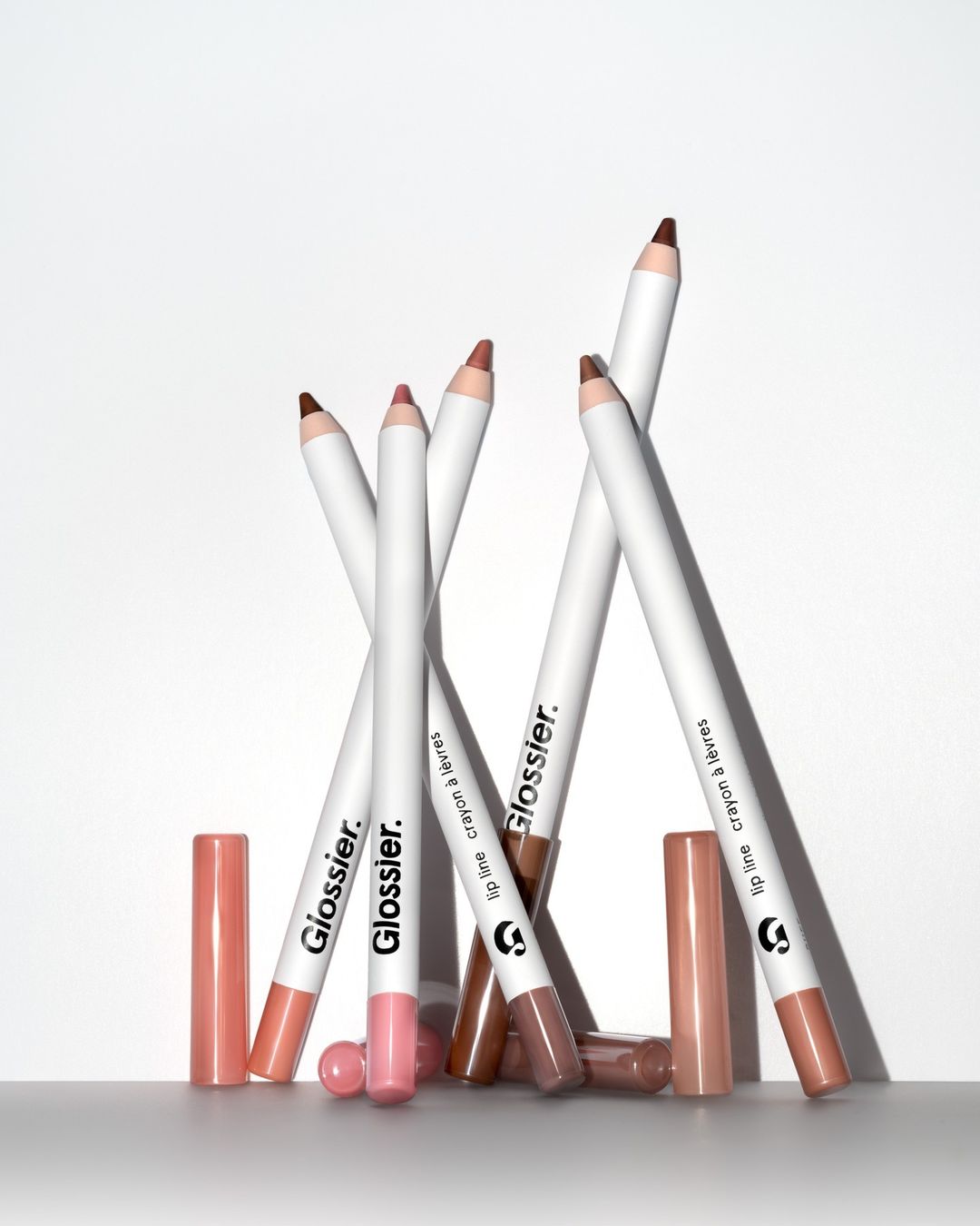 Close-up studio shot of multiple Glossier lip crayons with pink and brown shades, along with their caps, against a white background.
