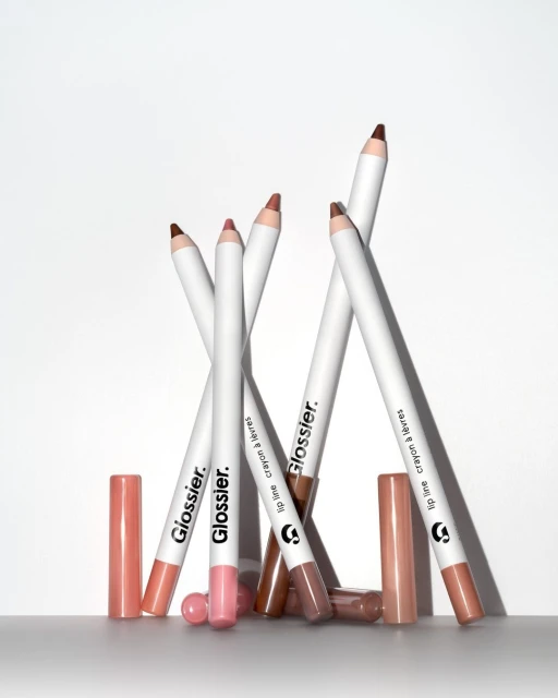 Close-up studio shot of multiple Glossier lip crayons with pink and brown shades, along with their caps, against a white background.