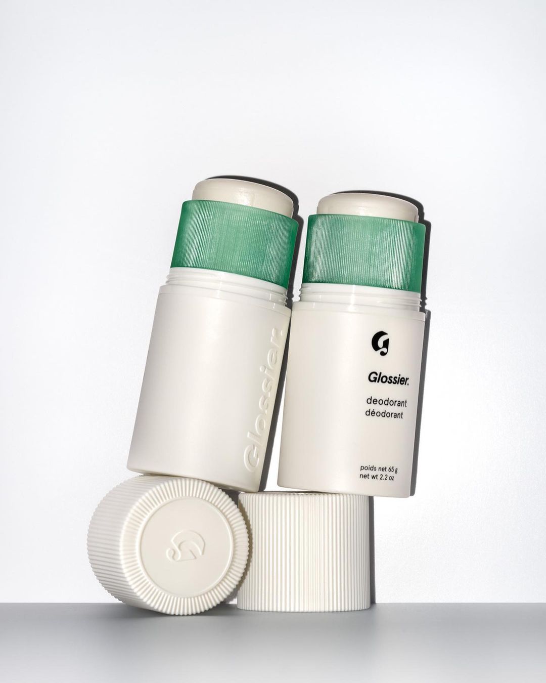 Close-up studio shot of two white Glossier deodorant sticks with green caps, one leaning against the other, and two white caps.